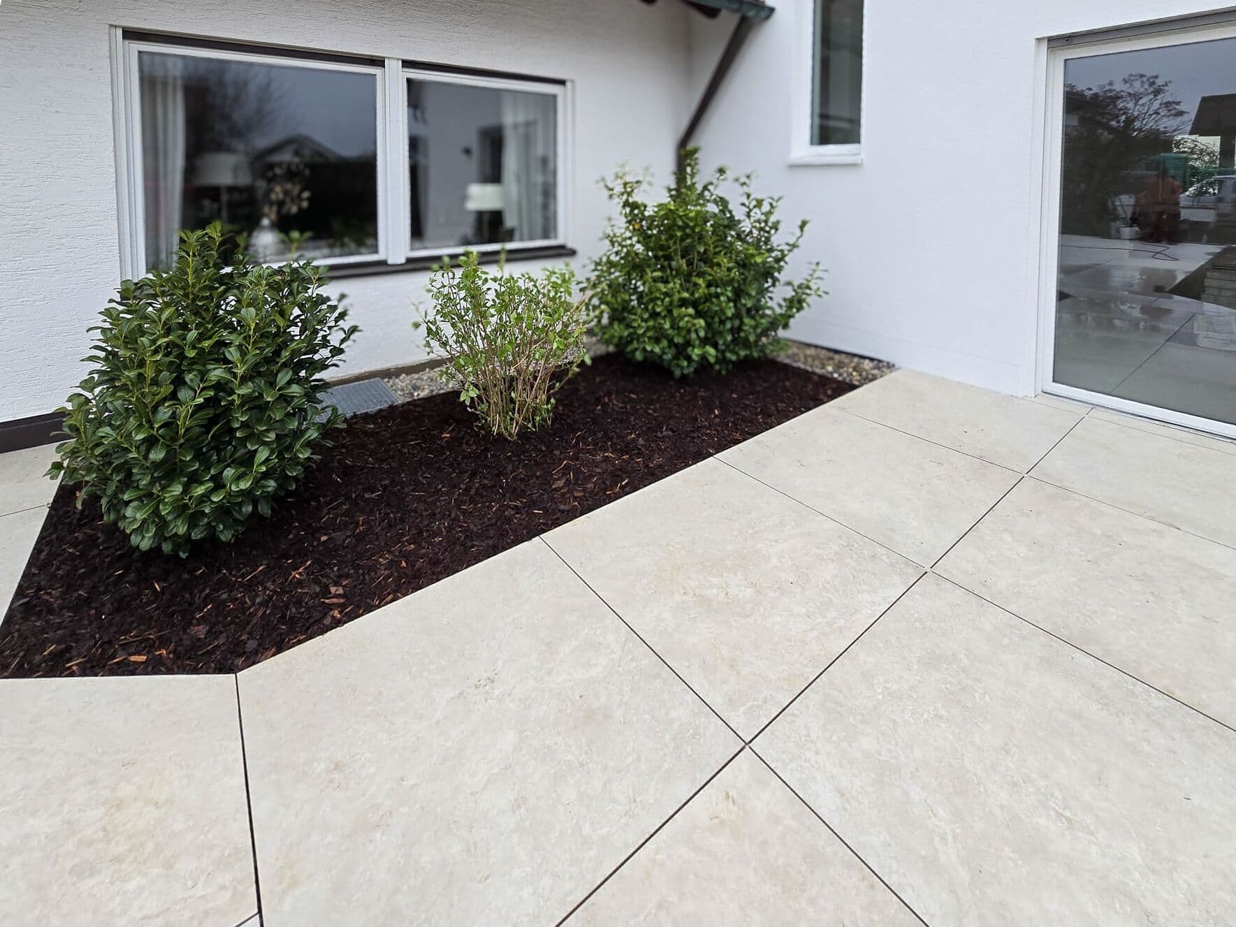 Helle Terrassenplatten am Haus mit angelegtem Beet und Sträuchern